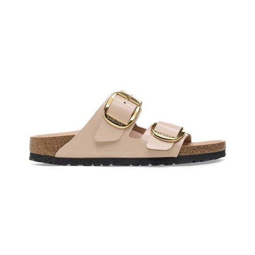 The BIRKENSTOCK ARIZONA BIG BUCKLE HIGH SHINE NEW BEIGE - WOMENS sandal features two wide straps made from high-quality natural leather, each adorned with a large gold buckle. The Birkenstock design includes a cork sole complemented by a black rubber bottom for comfort and durability.